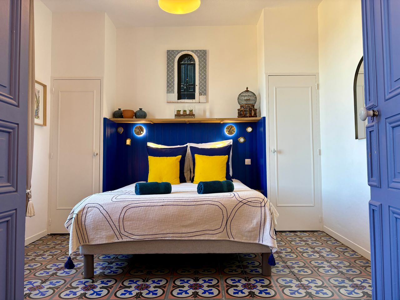 Intérieur d'une chambre, les draps du lit sont bleus, un tableau représentatn des fleurs est accroché au mur